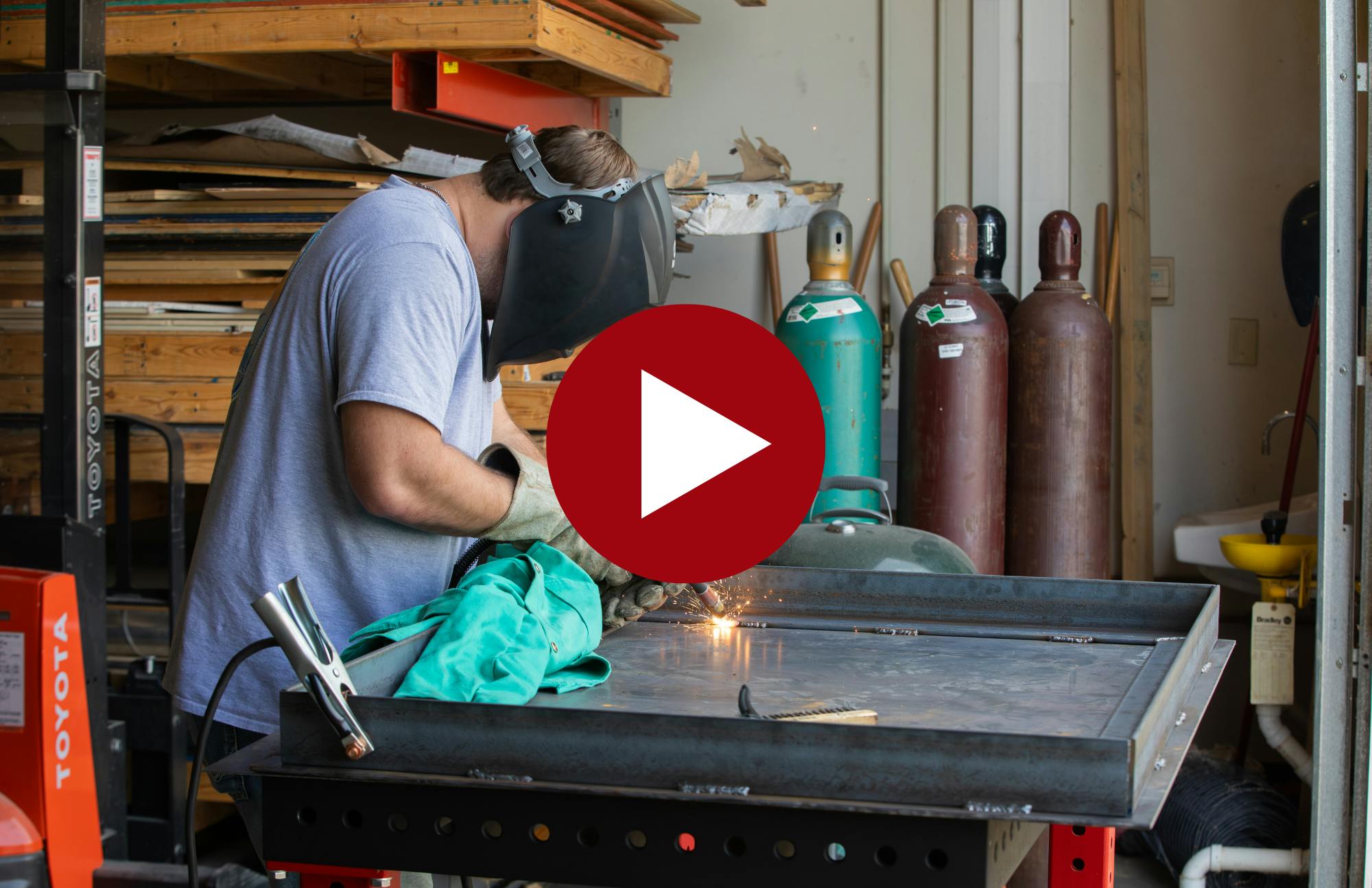 thumbnail image with a play button layered overtop an image of a student welding in the manufacturing lab  