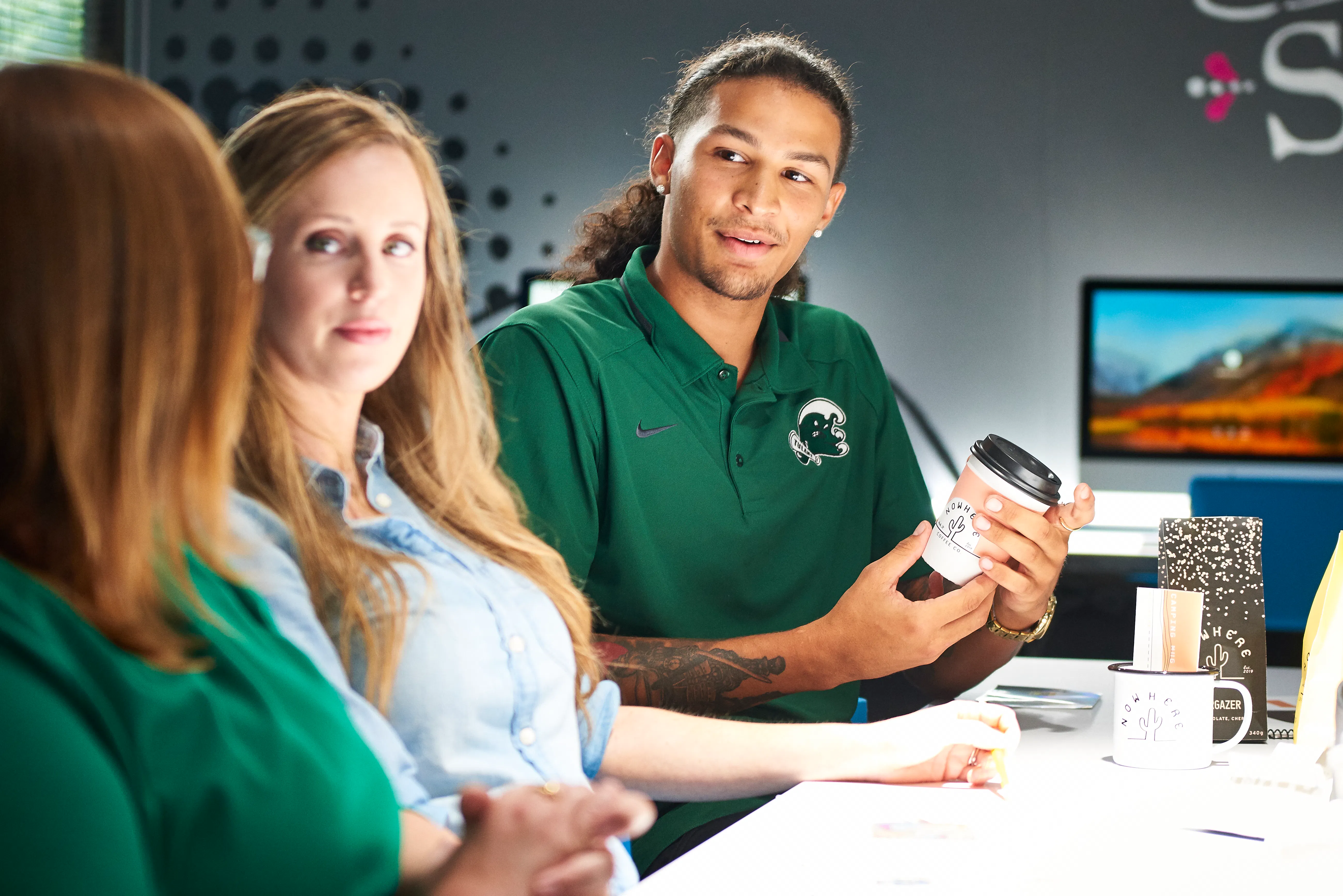 Three students in a campus study space in conversation; a young man in a green polo holds a coffee cup while two classmates listen around a white table.