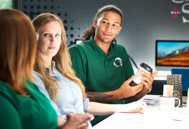 Three students in a campus study space in conversation; a young man in a green polo holds a coffee cup while two classmates listen around a white table.