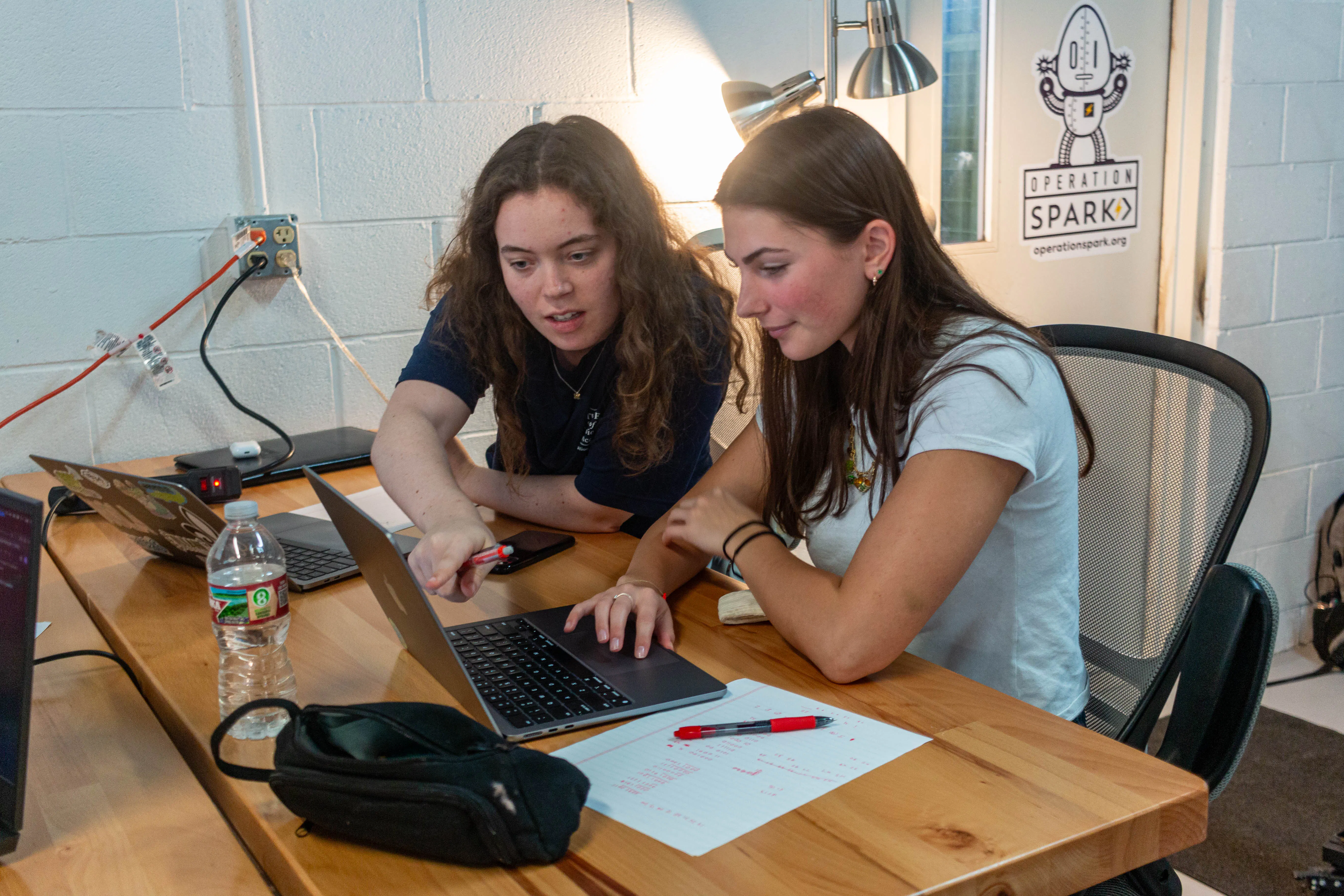 Two female students collaborating at a wooden table, one pointing at a laptop screen in a casual makerspace/tech workshop.