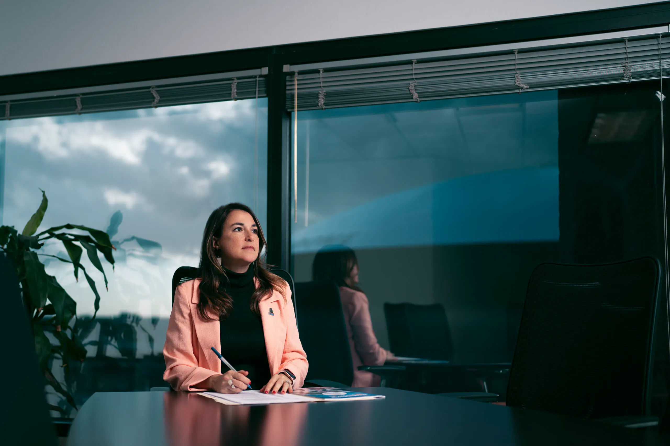 Professional woman in a boardroom looking out a window, pens and papers on the conference table.