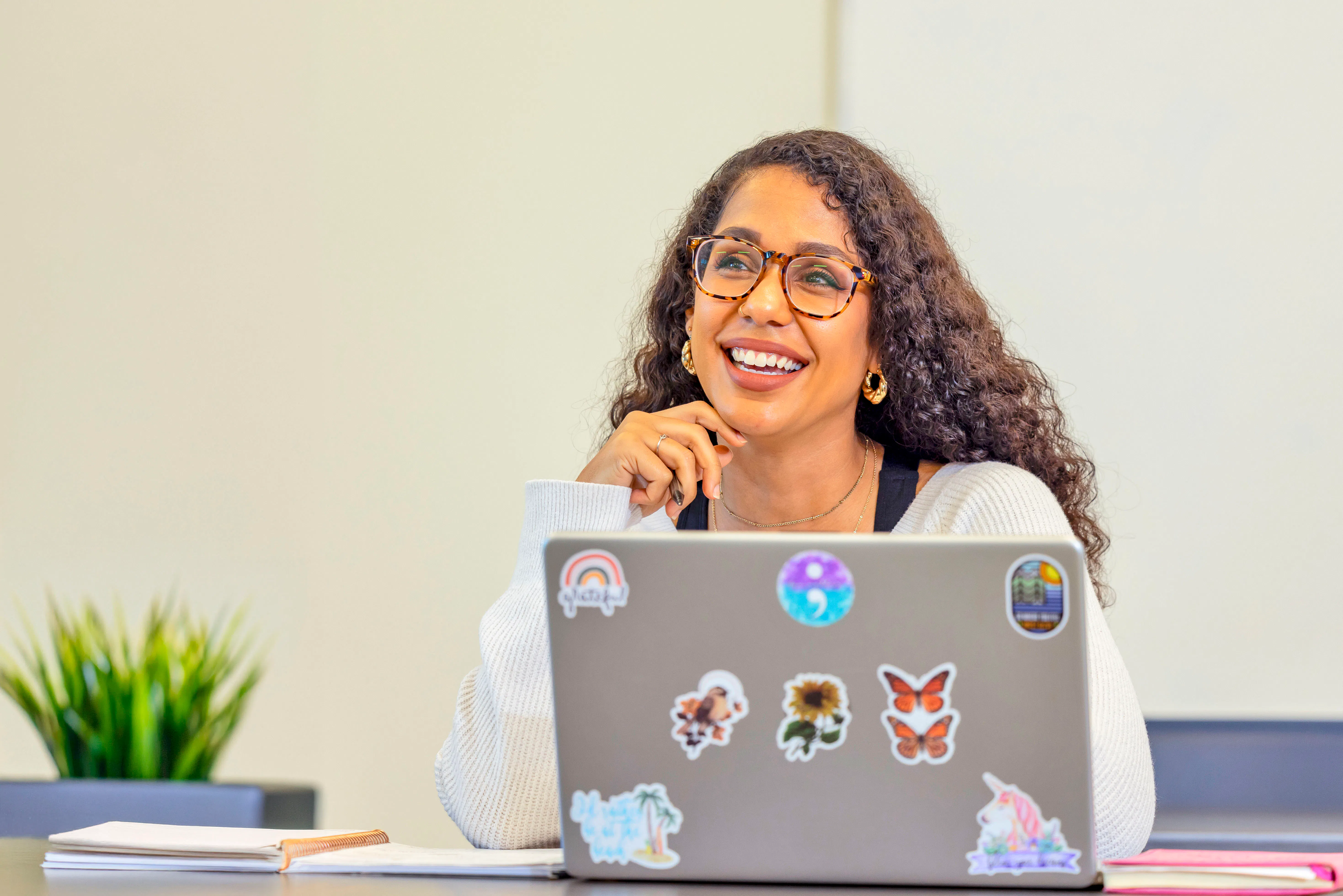 Smiling college student wearing glasses sits at a table and works on a laptop covered with colorful stickers. Papers and notebooks are visible on the table.