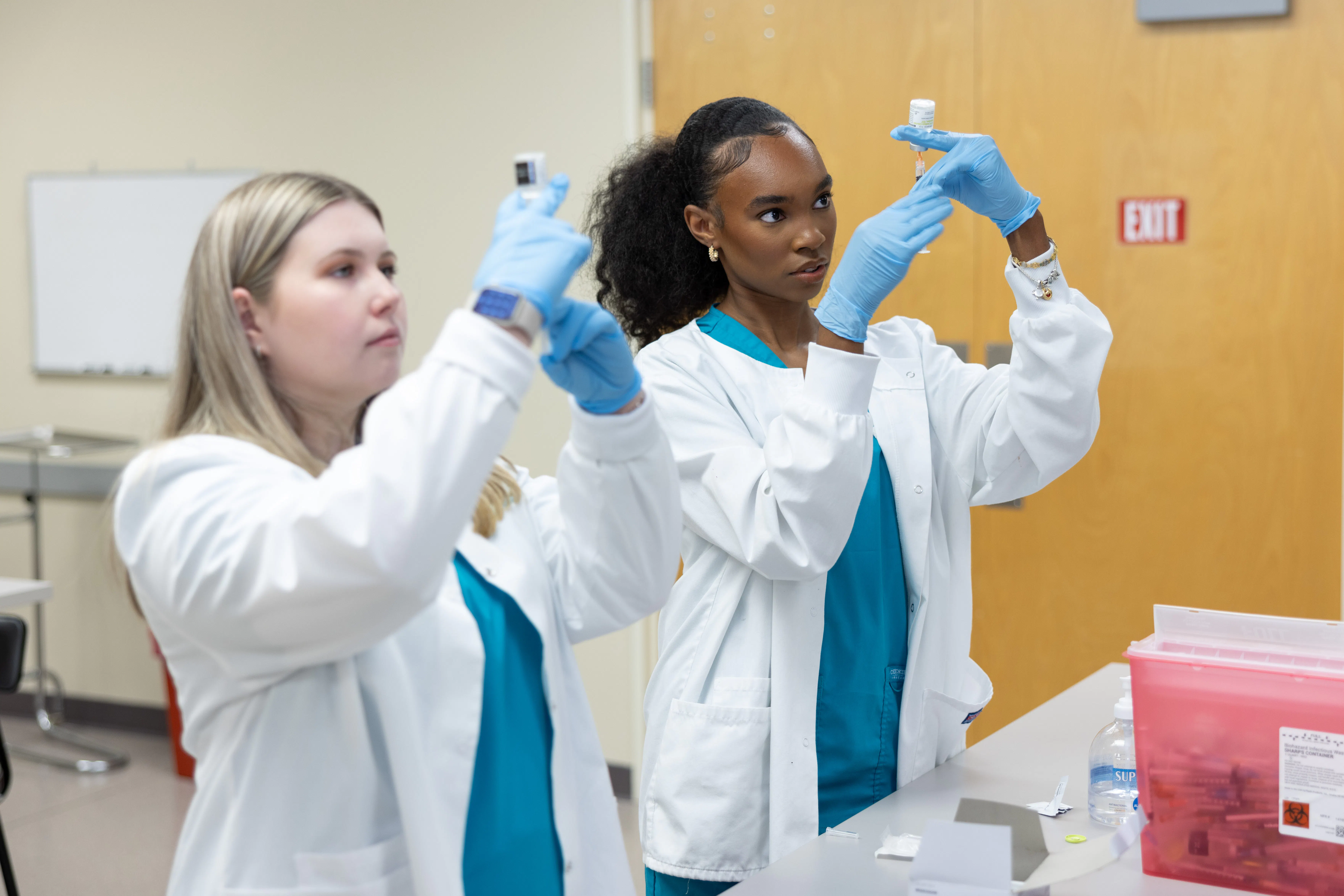 Wake Tech medical assisting students wearing lab coats and gloves prepare and examine syringes during a hands-on clinical skills training session in a classroom lab.