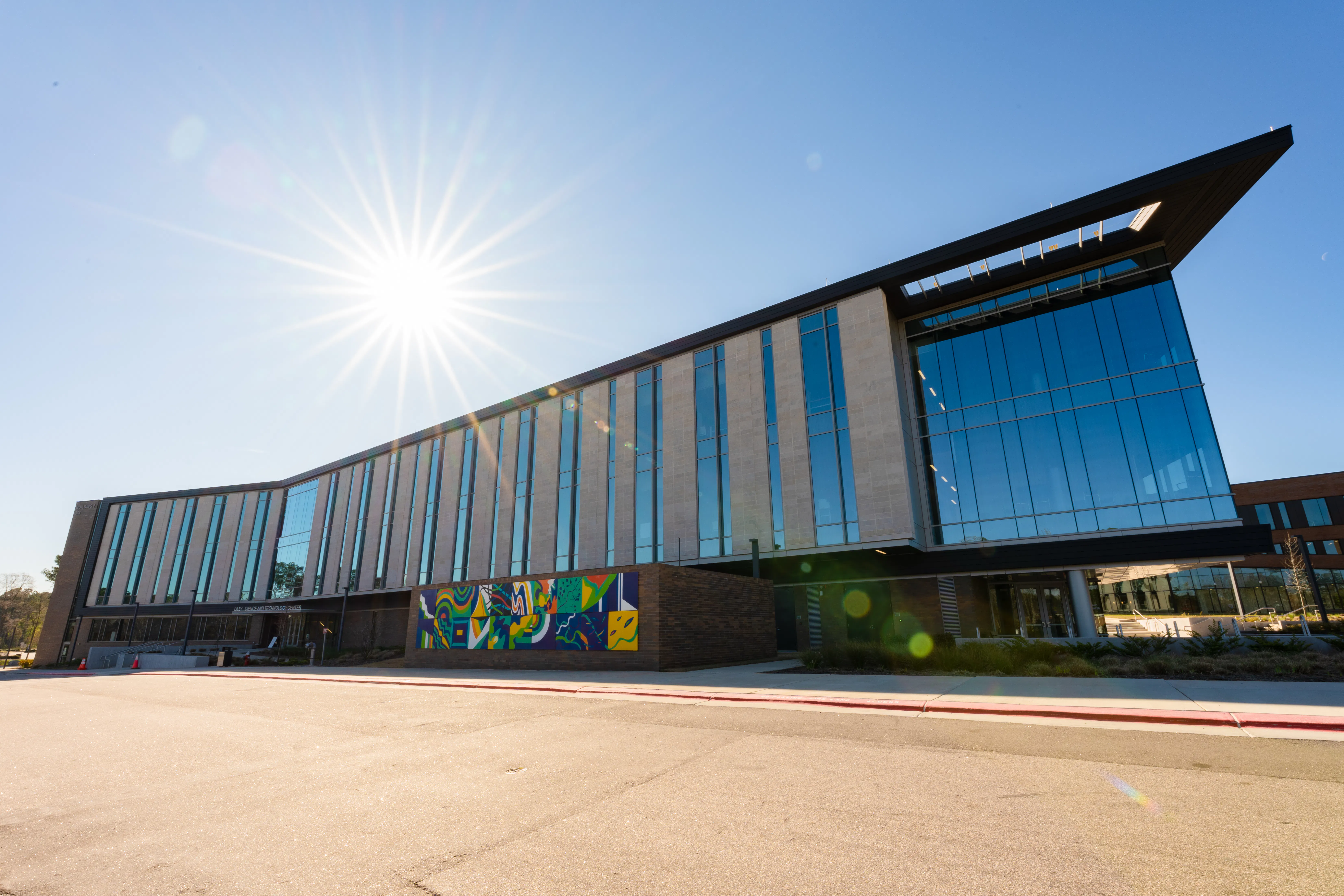 Wide exterior view of a contemporary Wake Tech campus building with tall vertical windows, a colorful mural on a brick wall near the entrance, and the sun shining brightly in a clear blue sky above.