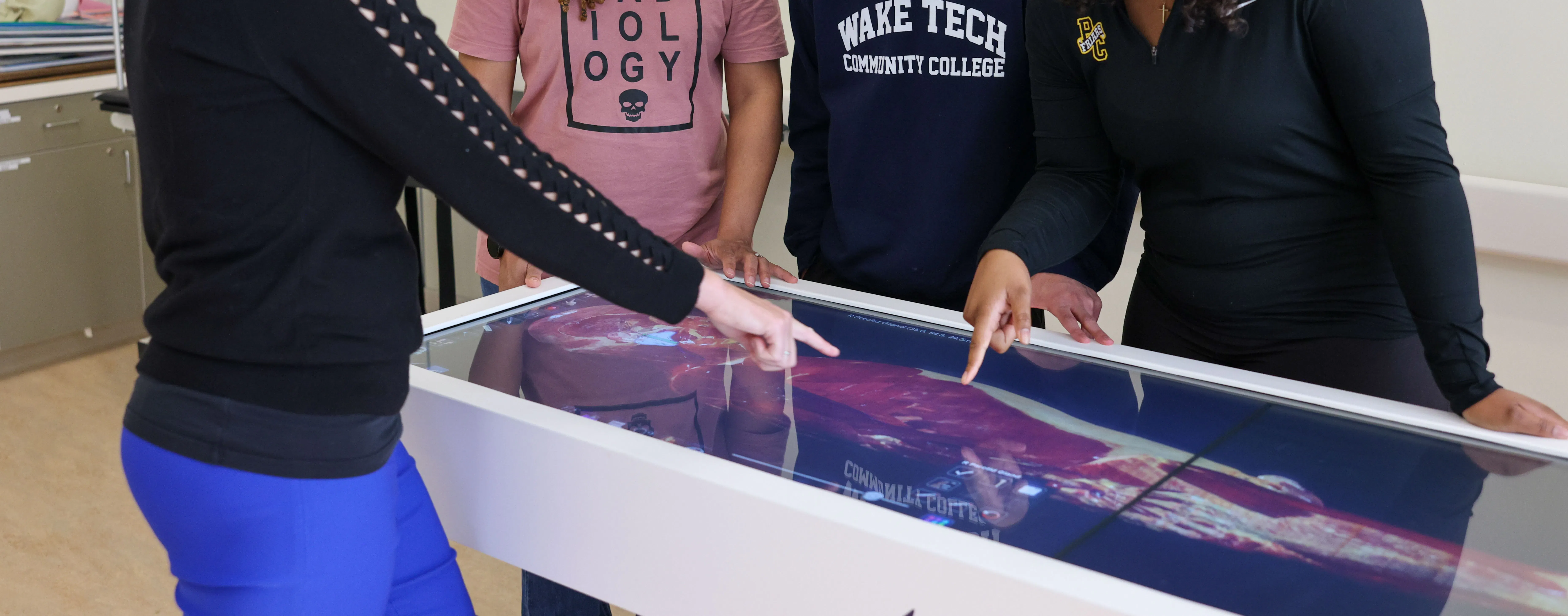 Wake Tech MRI students gather around an interactive anatomy table as an instructor points to detailed medical images, guiding students through imaging concepts in a classroom lab.