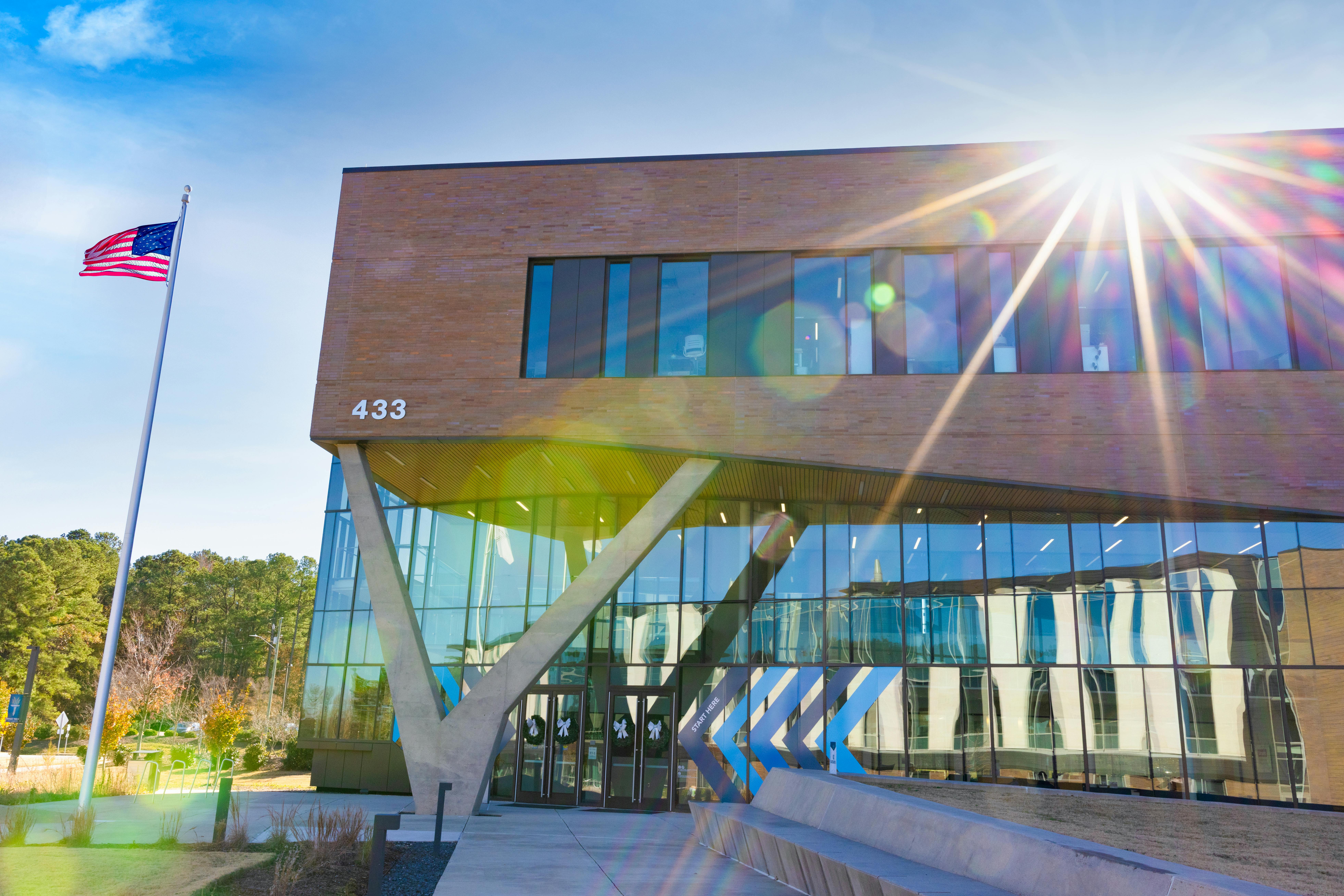 Modern Wake Tech campus building with large glass windows and angled concrete columns at the entrance, an American flag on a tall flagpole to the left, and bright sun rays creating a lens flare across the façade.