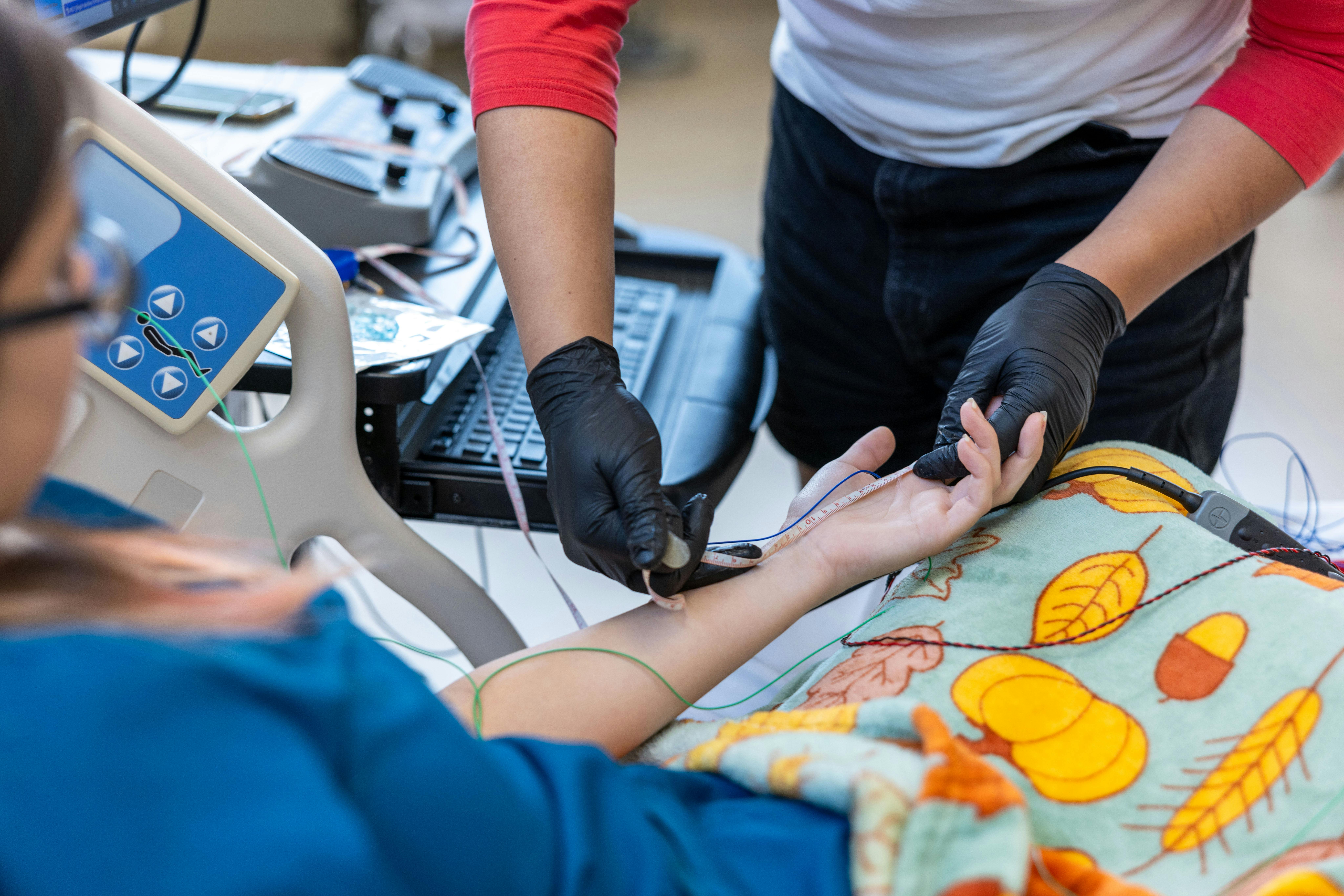 Wake Tech neurodiagnostic technology student wearing gloves places monitoring electrodes and measures a patient’s arm during a nerve conduction study in a clinical training lab, with diagnostic equipment and cables visible nearby.