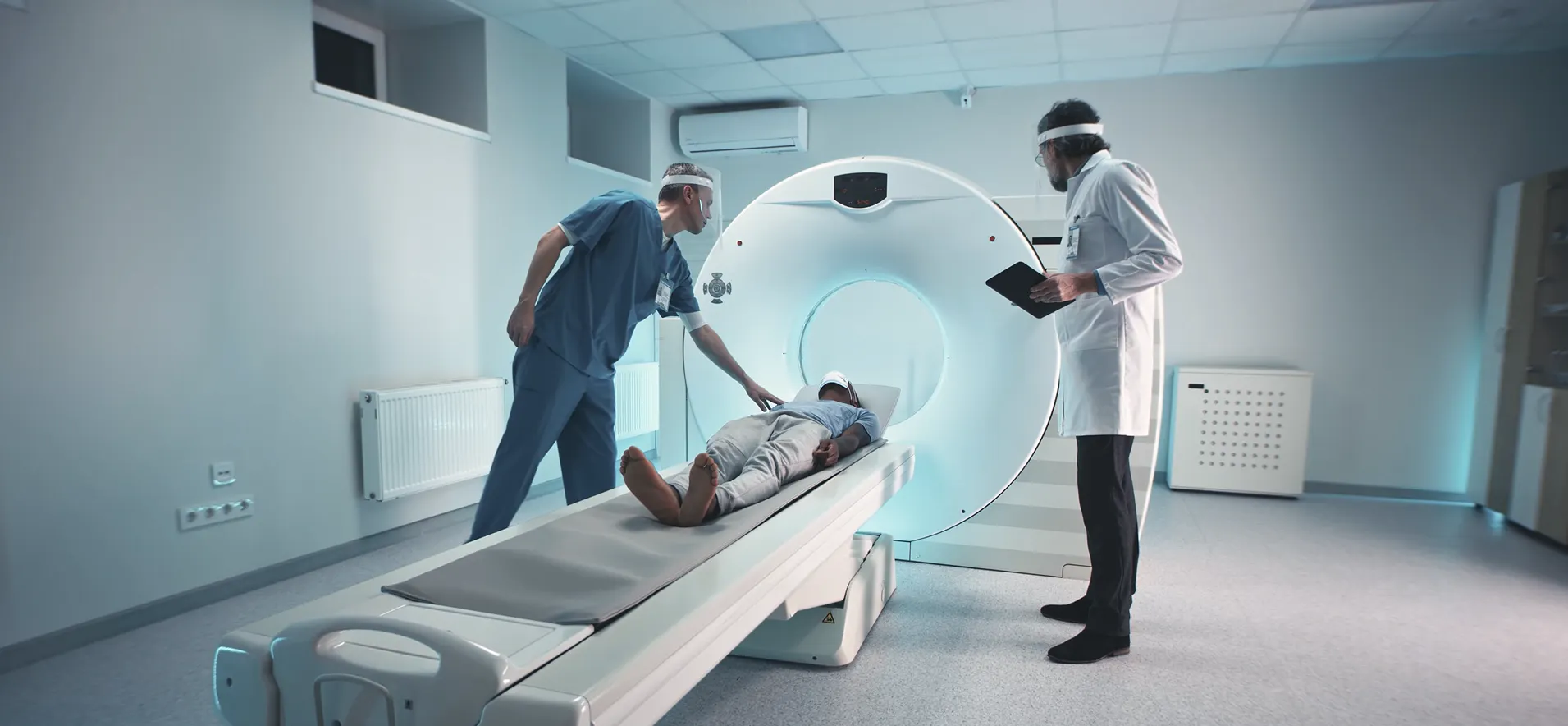 CT technology students observe and assist as a patient lies on a motorized table moving into a CT scanner, while an instructor reviews the procedure in a medical imaging lab.