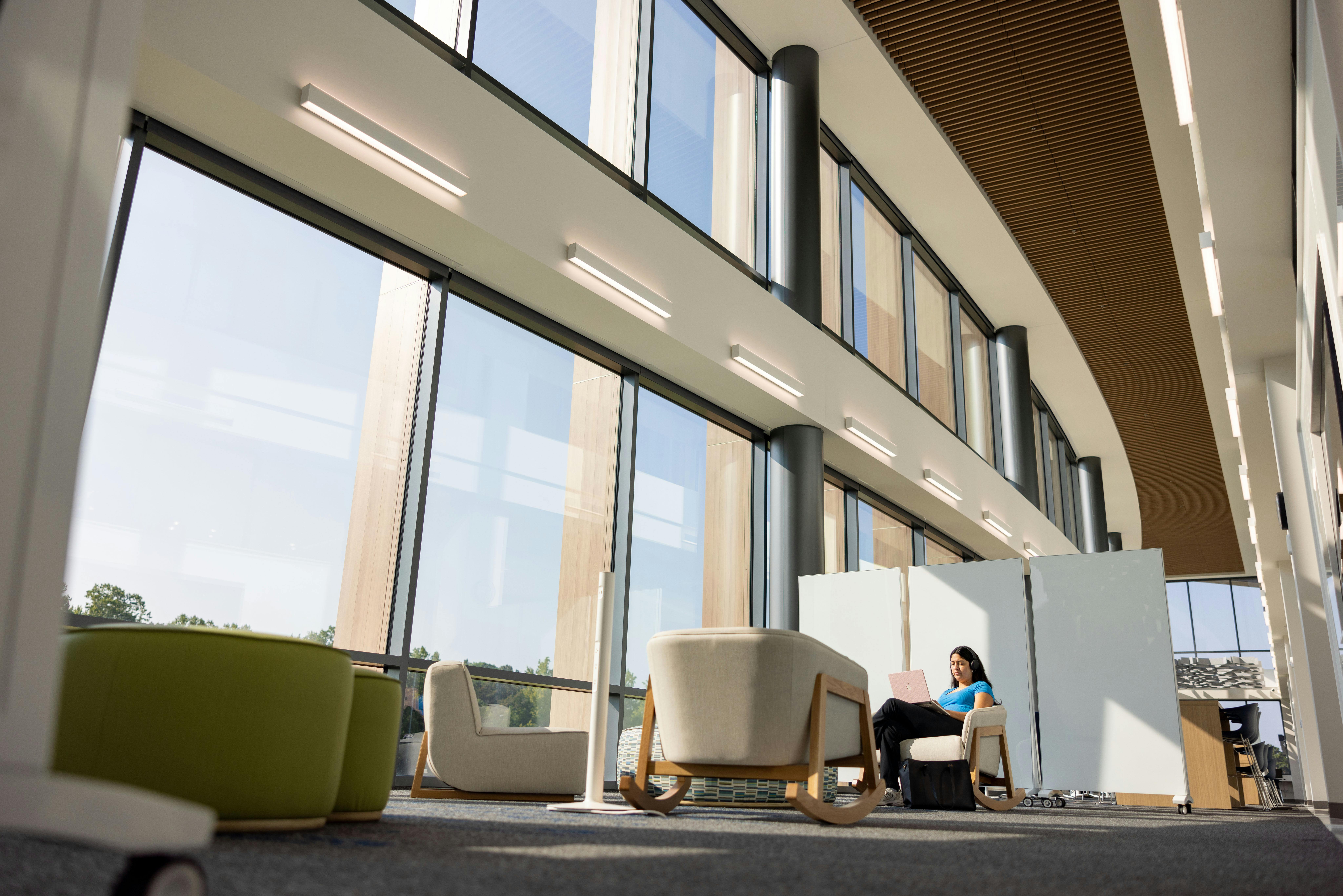 Interior of a Wake Tech campus building with floor-to-ceiling windows, wood slat ceiling panels, and soft lounge seating, where a student sits in a chair using a laptop in a quiet, sunlit study area.