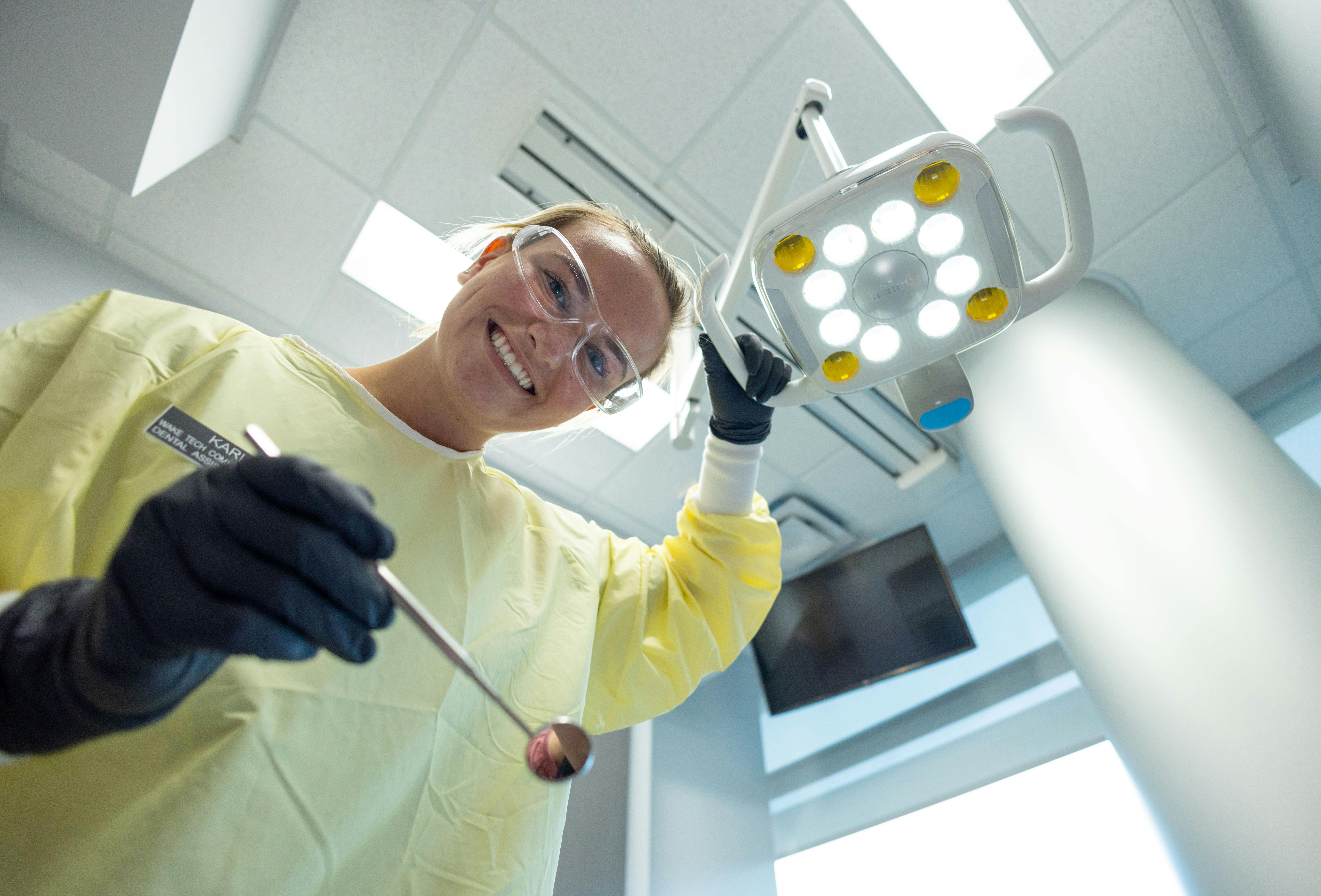 Wake Tech dental student wearing protective glasses, gloves, and a yellow clinical gown smiles while holding dental instruments under an overhead exam light in a dental training clinic.