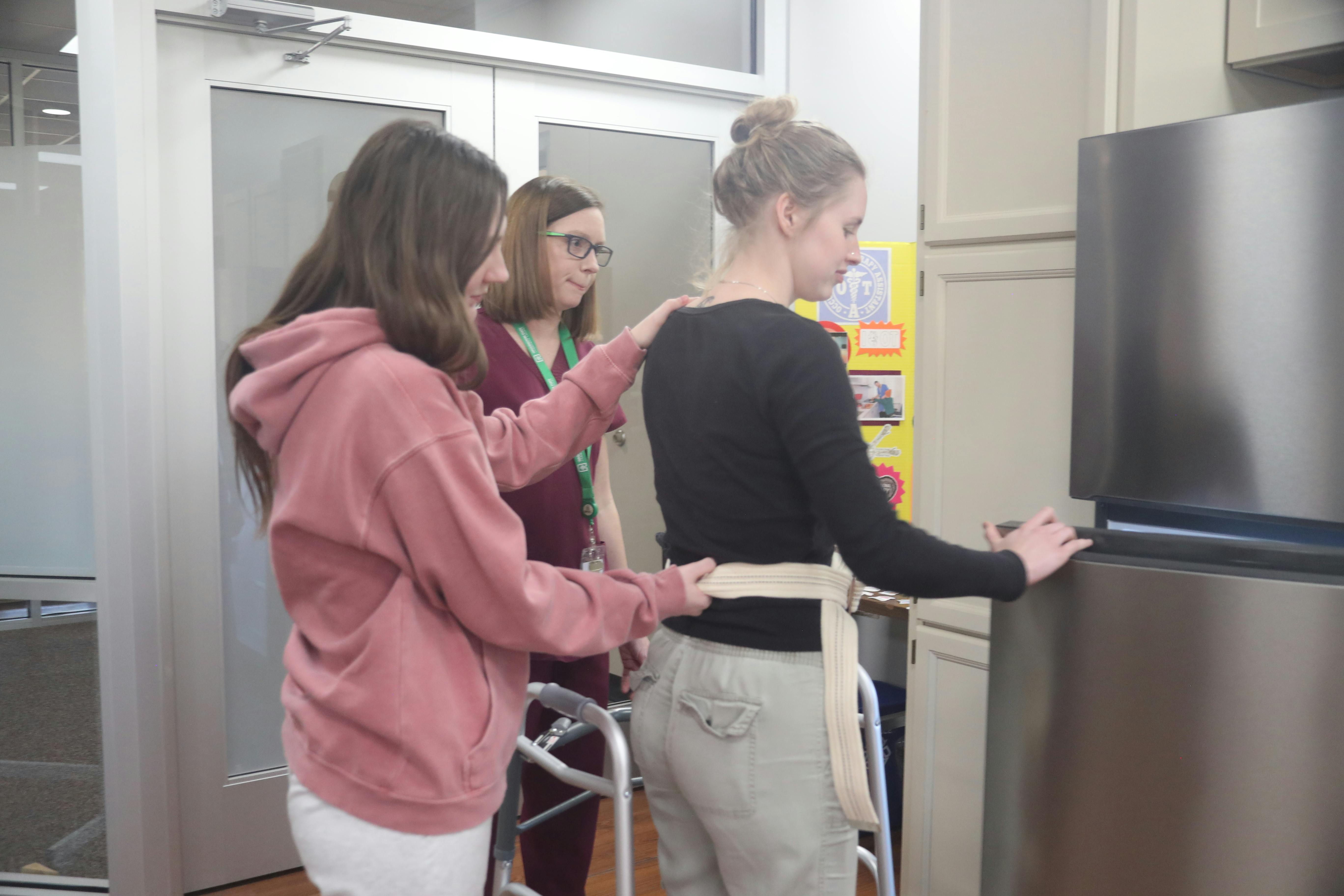 A professor observes as two students demonstrate proper technique for assisting a patient with using a refrigerator